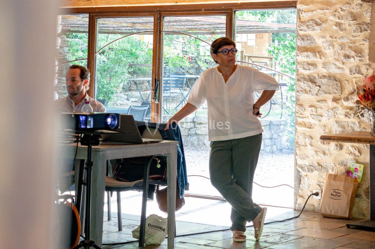 Une femme debout près d'une table avec un projecteur, dans une pièce lumineuse aux murs en pierre et grandes fenêtres.
