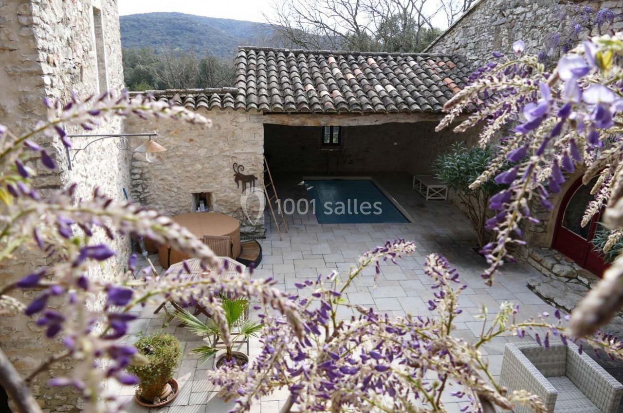 Cour en pierre avec piscine couverte, mobilier d'extérieur et glycine en fleurs, entourée de bâtiments en pierre.