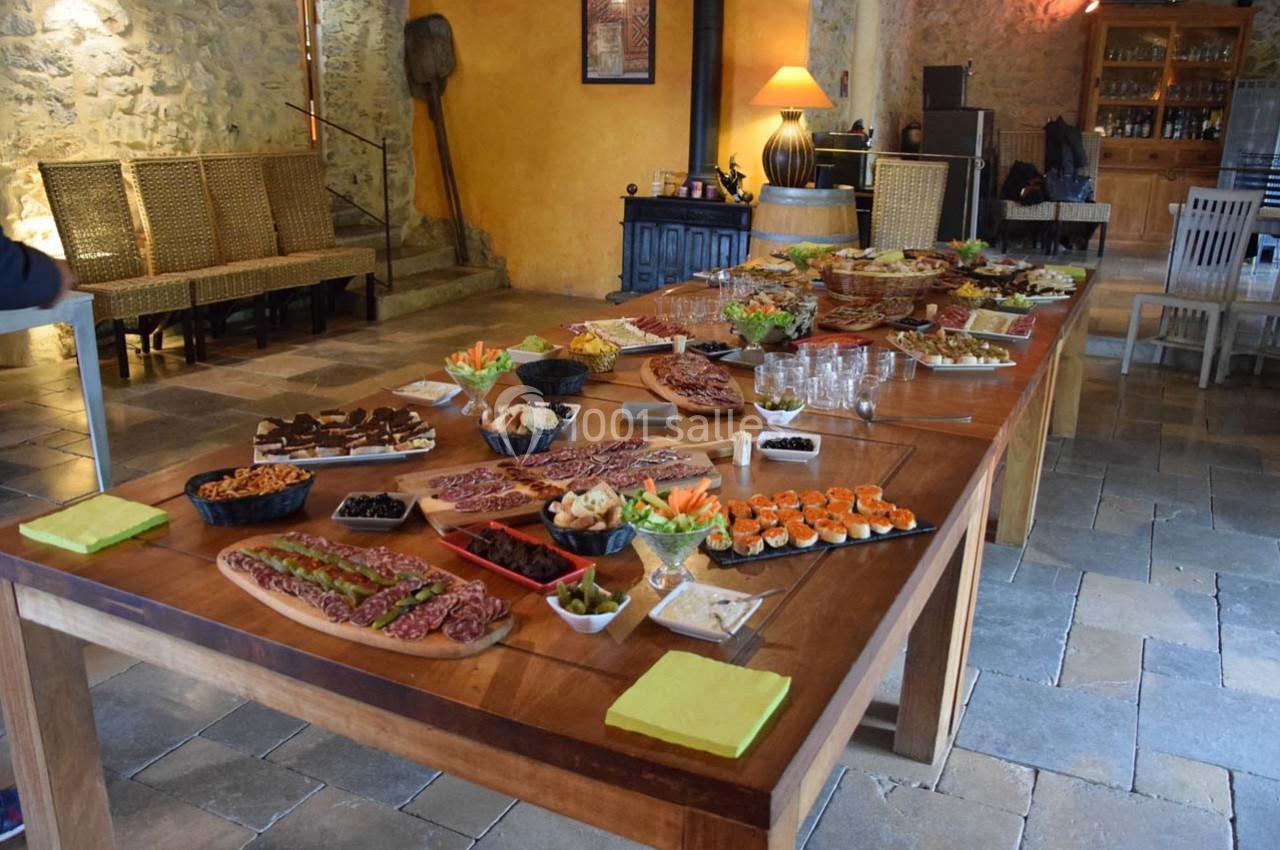 Grande table en bois garnie de plats variés, incluant charcuterie, légumes, desserts et apéritifs, dans une salle rustique.