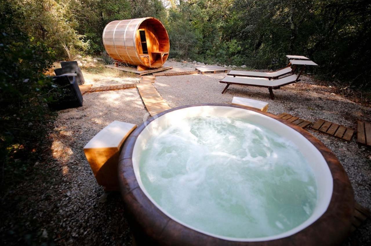 Bain à remous en extérieur avec sauna en bois et chaises longues, entourés de végétation naturelle.