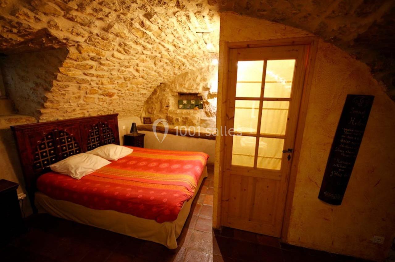 Chambre avec murs en pierre apparente, lit double avec couvre-lit rouge et porte en bois donnant sur une pièce éclairée.