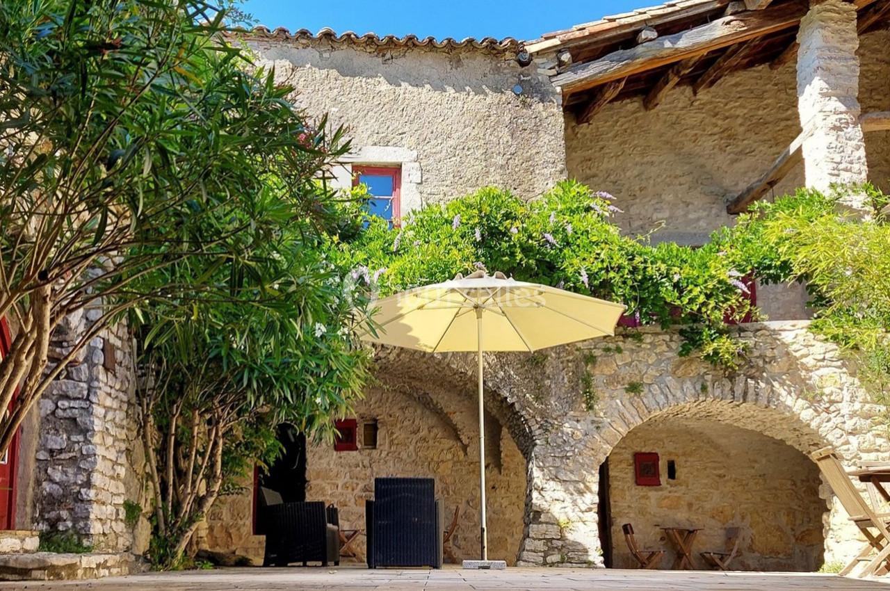 Cour ensoleillée avec murs en pierre, végétation grimpante, parasol beige et mobilier d'extérieur en osier noir.