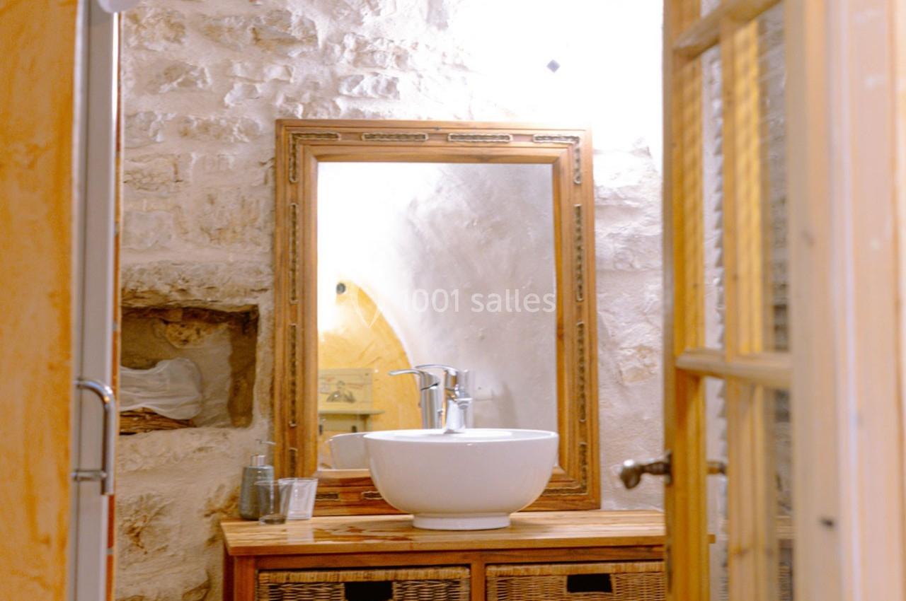 Lavabo blanc posé sur un meuble en bois avec miroir encadré, dans une salle de bain aux murs en pierre apparente.