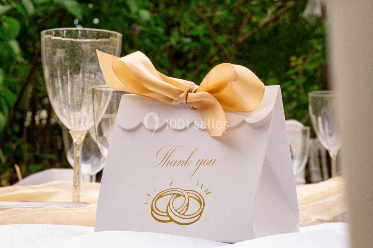 Boîte cadeau blanche avec un ruban doré, posée sur une table décorée, entourée de verres et de verdure en arrière-plan.