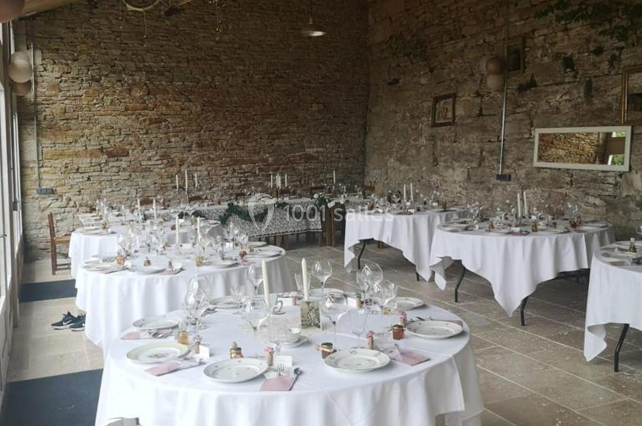 Salle de réception en pierre avec tables rondes dressées pour un événement, décorées de nappes blanches et bougies.