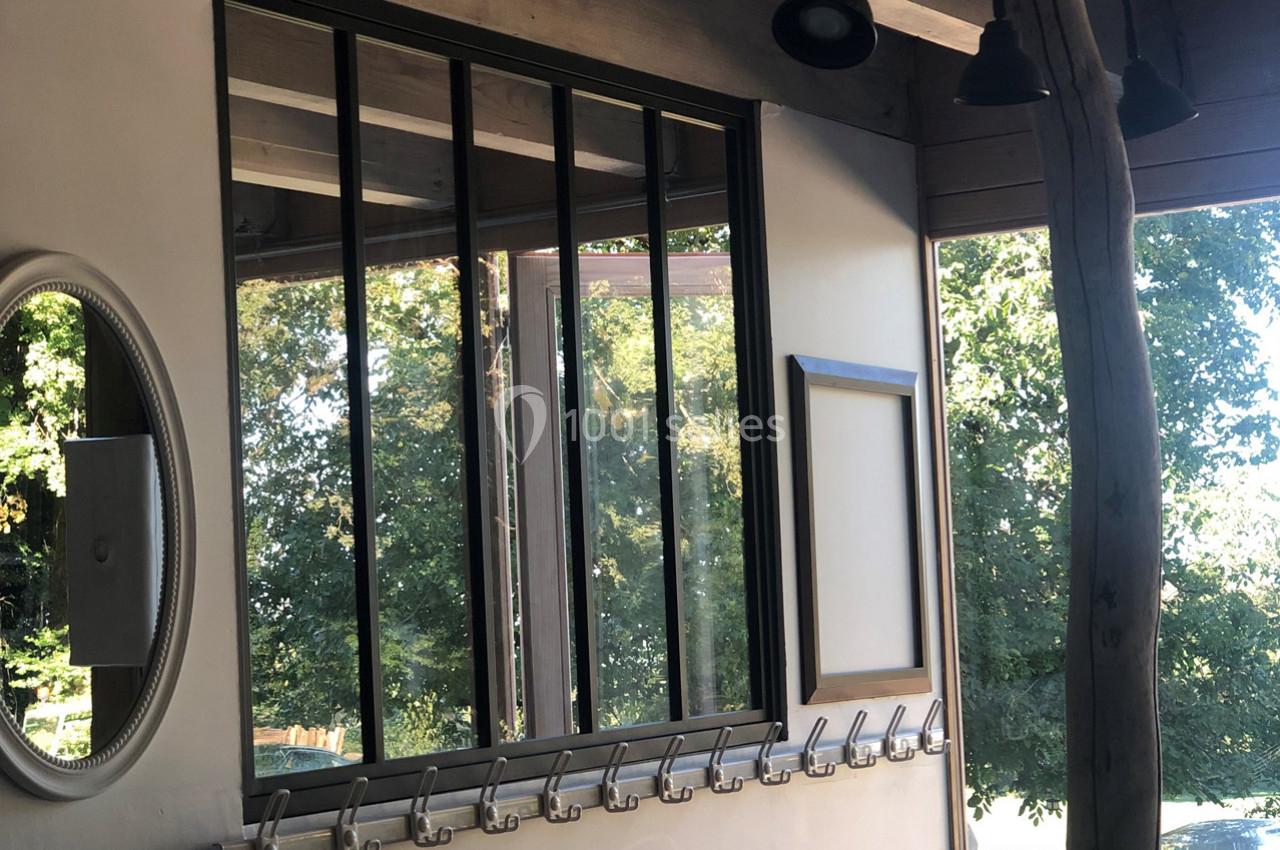 Mur intérieur avec verrière noire, miroir rond, patères métalliques et vue sur des arbres à travers une fenêtre.