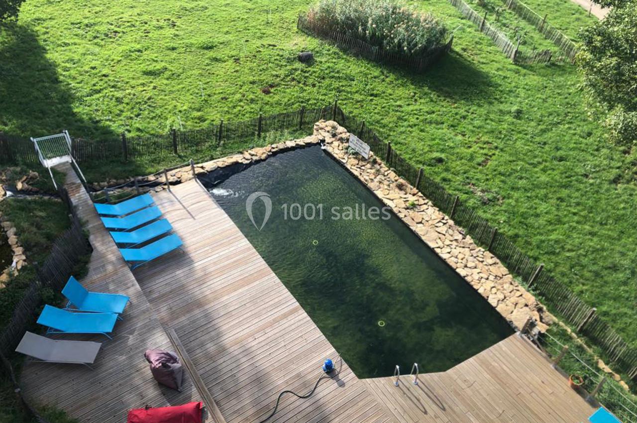 Vue aérienne d'une piscine naturelle entourée d'une terrasse en bois, avec des transats bleus et un jardin verdoyant.