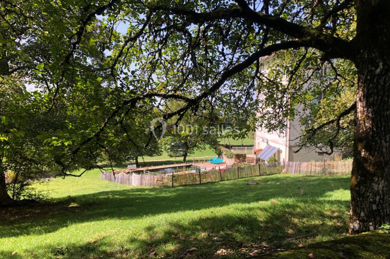 Vue d'un jardin verdoyant avec une piscine clôturée, un bâtiment blanc en arrière-plan et des arbres au premier plan.