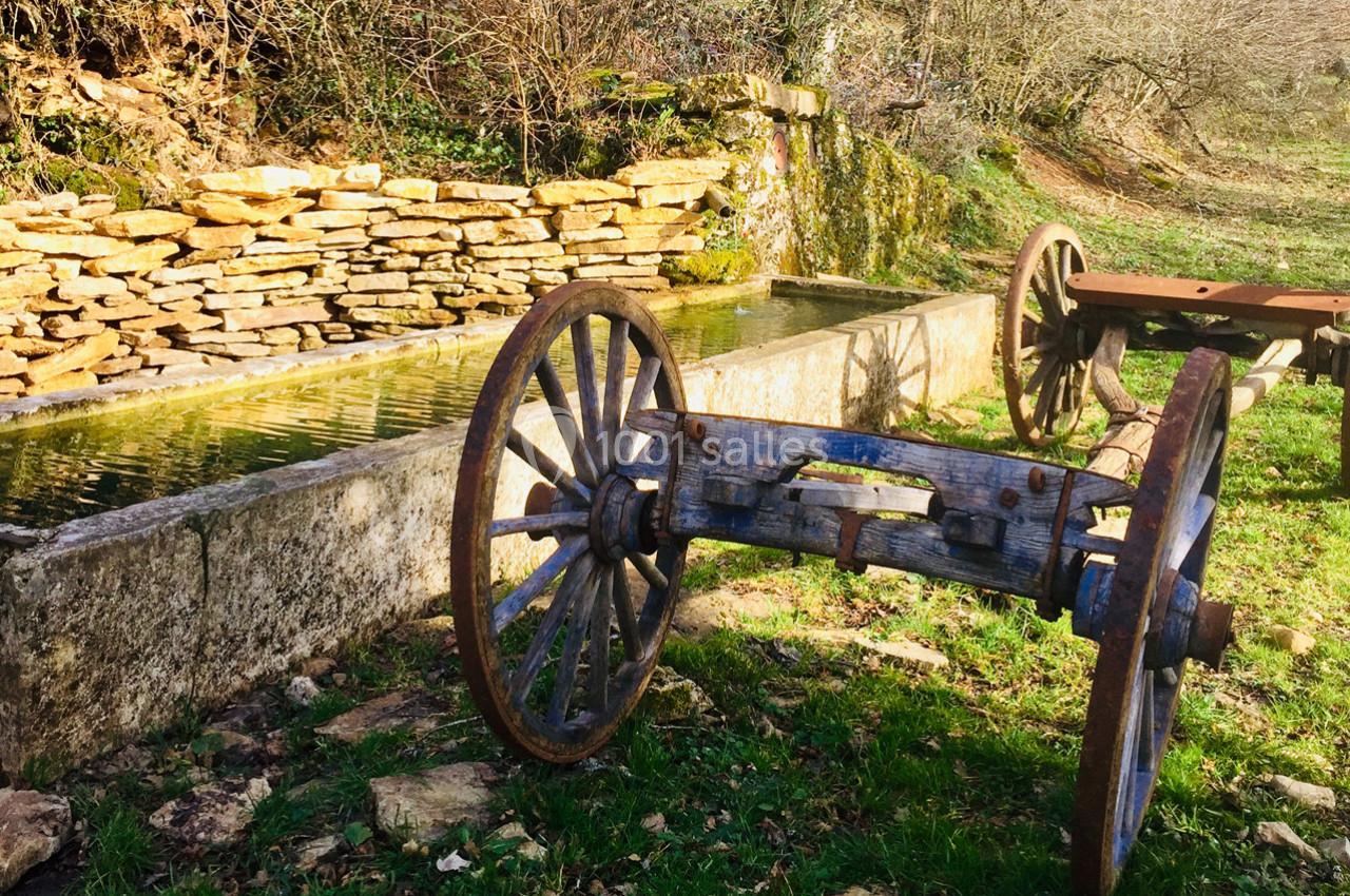 Vieille charrette en bois abandonnée près d'un abreuvoir en pierre entouré de végétation et d'un mur en pierres sèches.