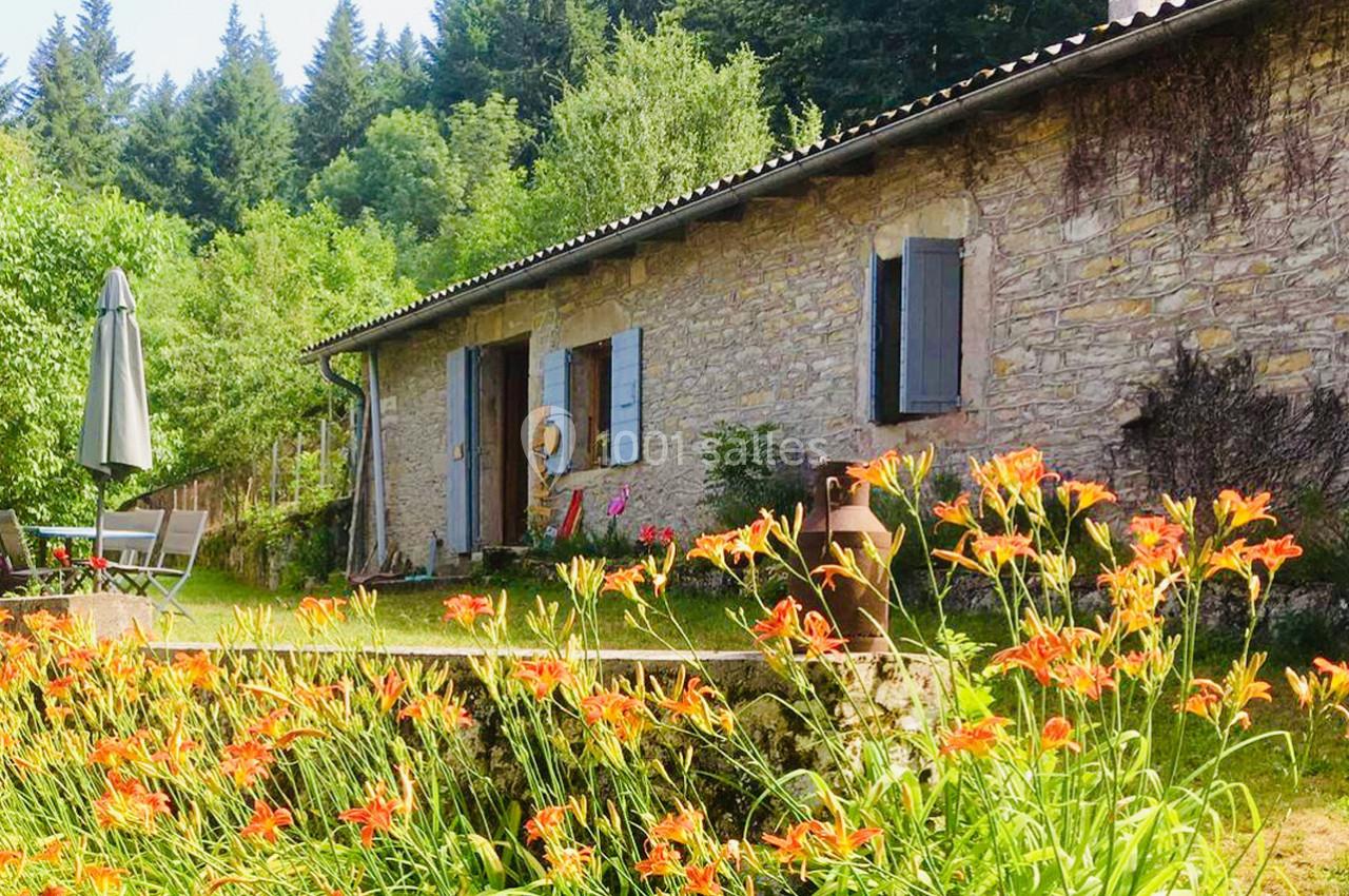 Maison en pierre avec volets bleus, entourée de fleurs orange et de verdure, située près d'une forêt.