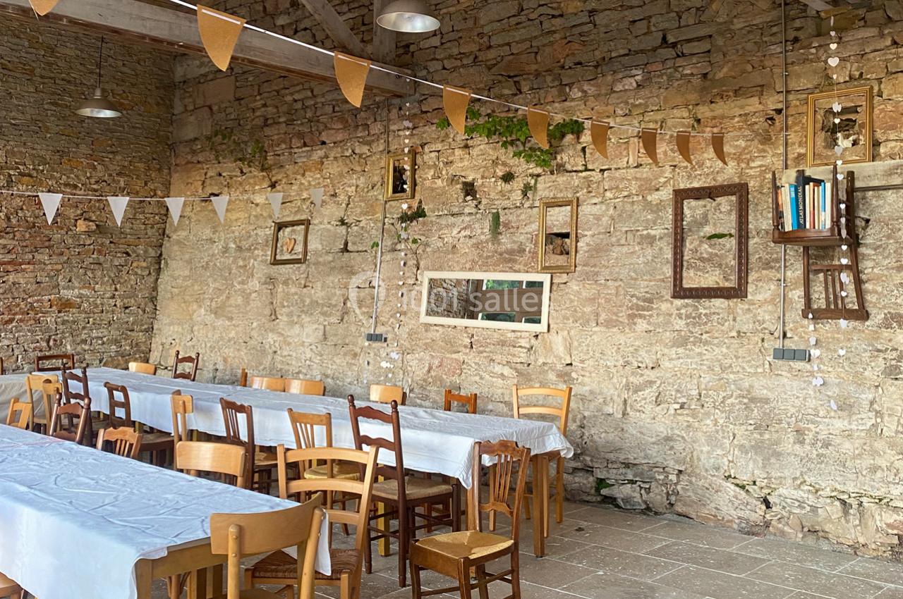 Salle rustique avec murs en pierre, tables alignées couvertes de nappes blanches et décorations suspendues.