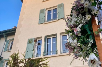 Façade d'une maison beige à volets verts, reflétée dans un bassin entouré de pierres et de végétation.