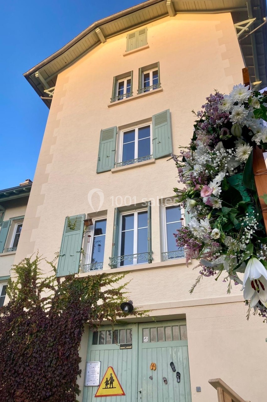 Façade d'une maison beige à volets verts, ornée de fleurs et de lierre, avec un panneau de signalisation devant.