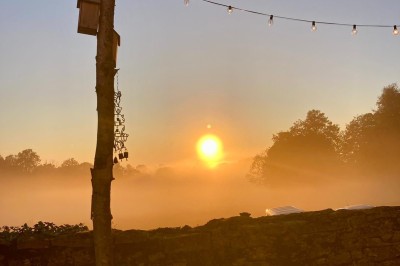 Façade d'une maison beige à volets verts, reflétée dans un bassin entouré de pierres et de végétation.