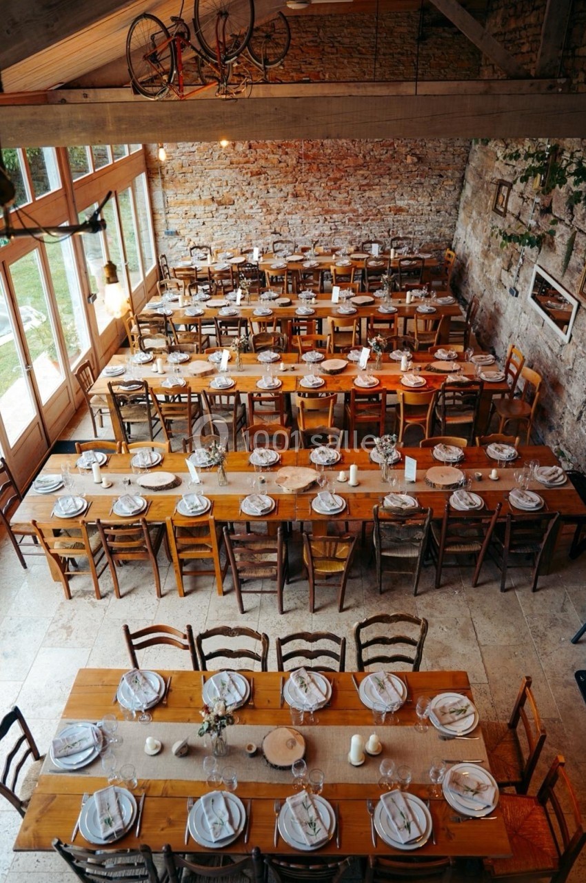 Grande salle avec de longues tables en bois dressées pour un repas, éclairée par des baies vitrées et des murs en pierre.