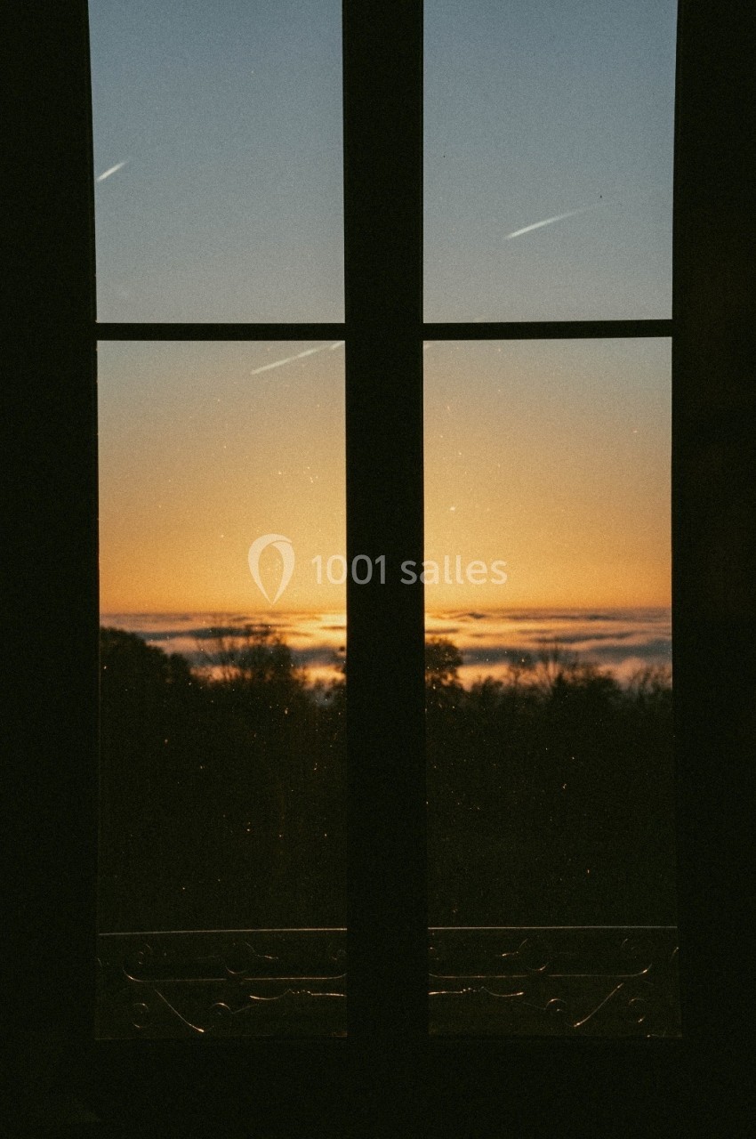 Vue d'un coucher de soleil à travers une fenêtre, avec des arbres en contre-jour et des traînées d'avion dans le ciel.