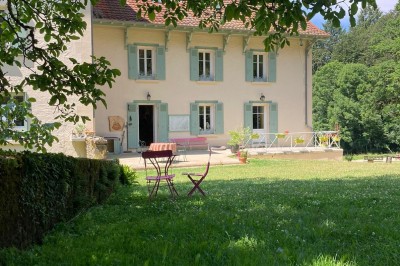 Façade d'une maison beige à volets verts, reflétée dans un bassin entouré de pierres et de végétation.