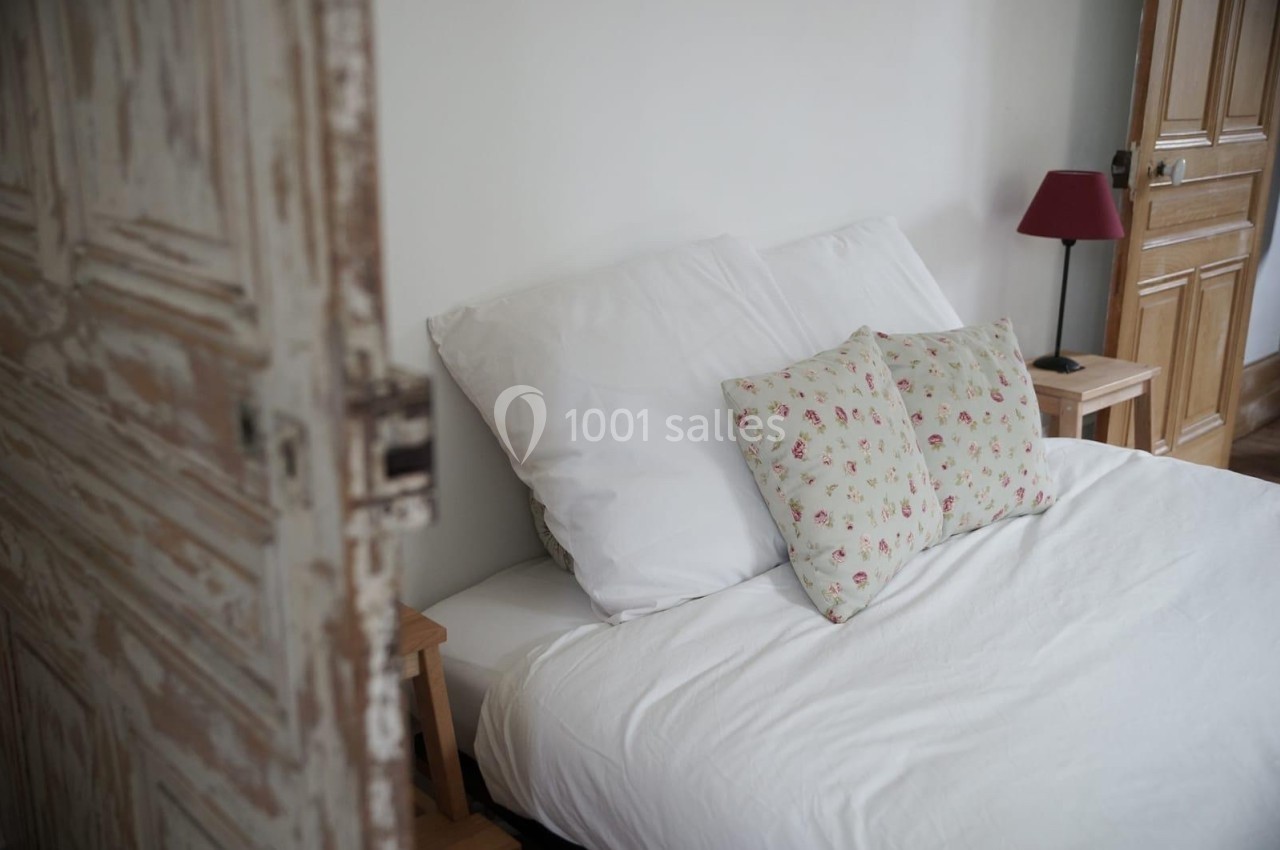 Lit avec draps blancs et coussins à motifs floraux, dans une chambre lumineuse avec une porte en bois vieilli.
