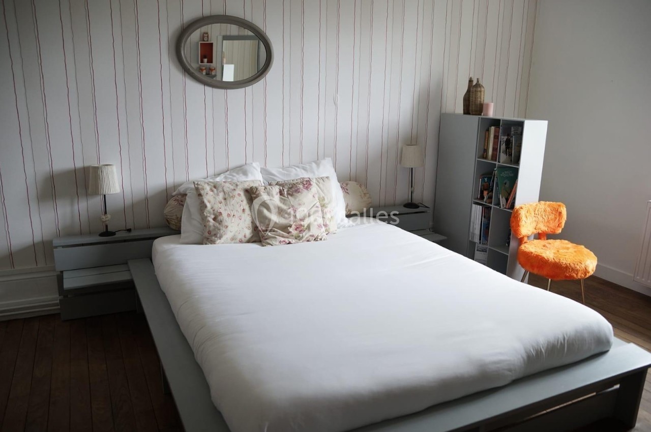 Lit double avec draps blancs, coussins à motifs floraux, étagère avec livres et chaise orange dans une chambre lumineuse.