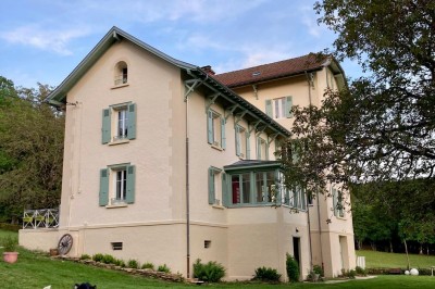 Façade d'une maison beige à volets verts, reflétée dans un bassin entouré de pierres et de végétation.