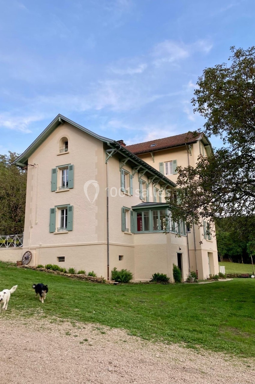 Grande maison beige à volets verts entourée de pelouse, avec deux chiens au premier plan et un arbre à droite.