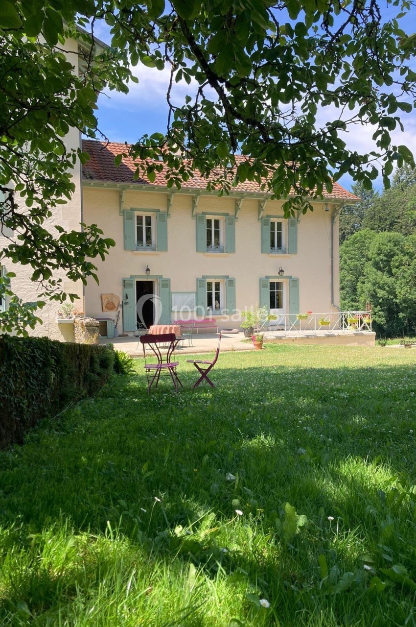 Façade d'une maison avec volets verts, terrasse aménagée et jardin verdoyant sous un ciel bleu.