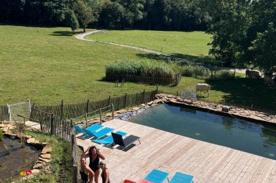 Façade d'une maison beige à volets verts, reflétée dans un bassin entouré de pierres et de végétation.