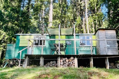Façade d'une maison beige à volets verts, reflétée dans un bassin entouré de pierres et de végétation.