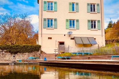 Façade d'une maison beige à volets verts, reflétée dans un bassin entouré de pierres et de végétation.