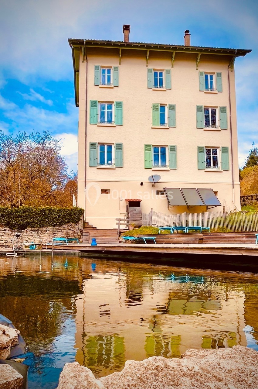 Façade d'une maison beige à volets verts, reflétée dans un bassin entouré de pierres et de végétation.