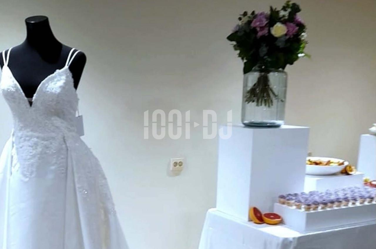Robe de mariée blanche sur mannequin, bouquet de fleurs dans un vase et table avec desserts en arrière-plan.