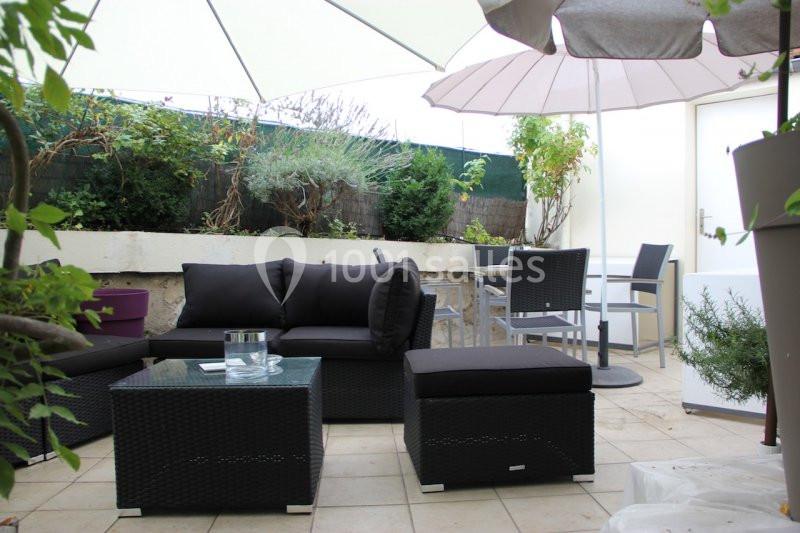 Salon de jardin avec mobilier en résine tressée, parasols ouverts et végétation en arrière-plan sur une terrasse carrelée.