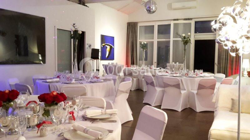 Salle de réception décorée avec des tables rondes dressées, nappes blanches, fleurs rouges et éclairage tamisé.