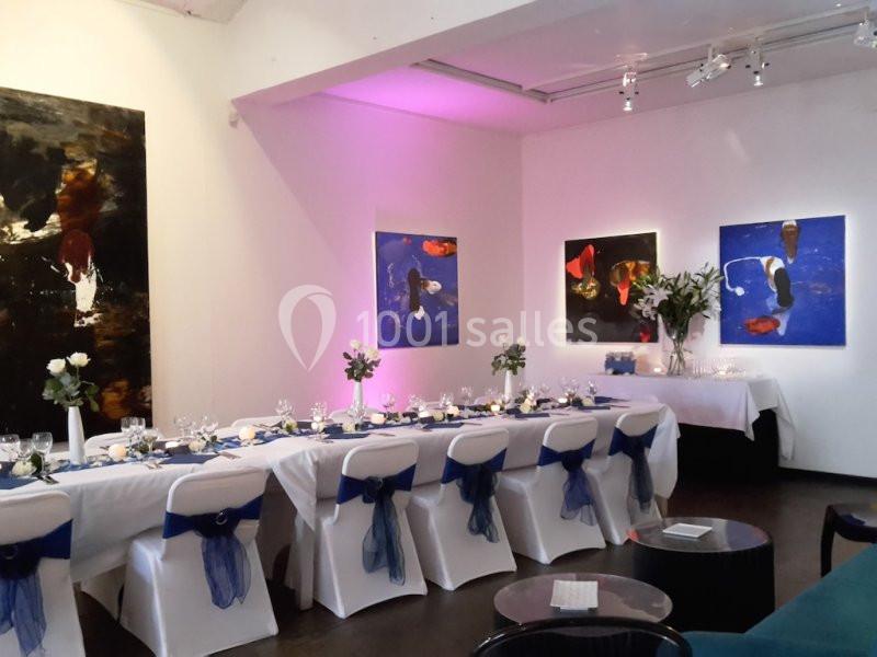 Salle de réception décorée avec des tables dressées, des chaises ornées de nœuds bleus et des tableaux colorés aux murs.