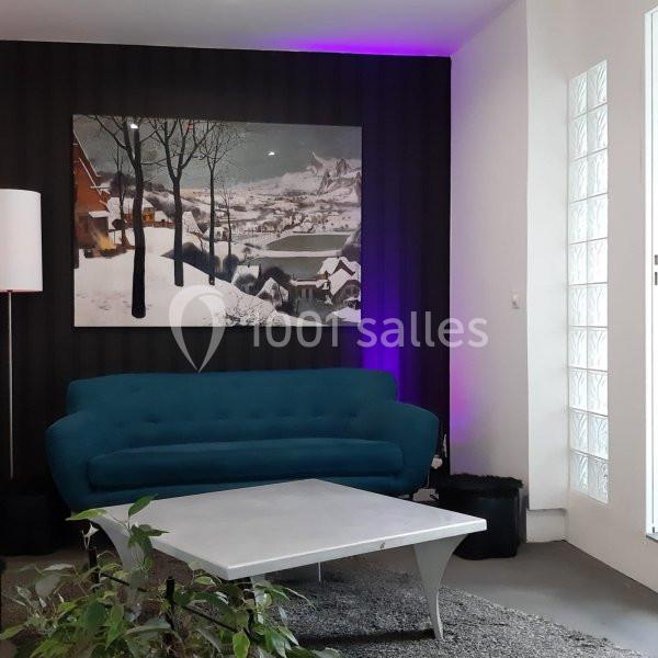 Salon moderne avec canapé bleu, table basse blanche, tableau de paysage enneigé et éclairage violet en arrière-plan.
