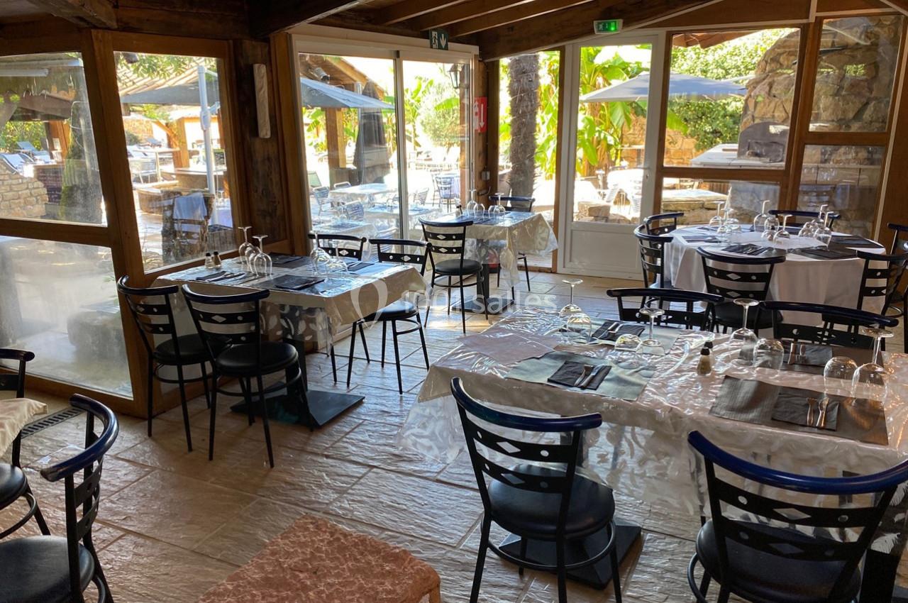 Salle de restaurant lumineuse avec tables dressées, chaises noires et vue sur une terrasse extérieure arborée.