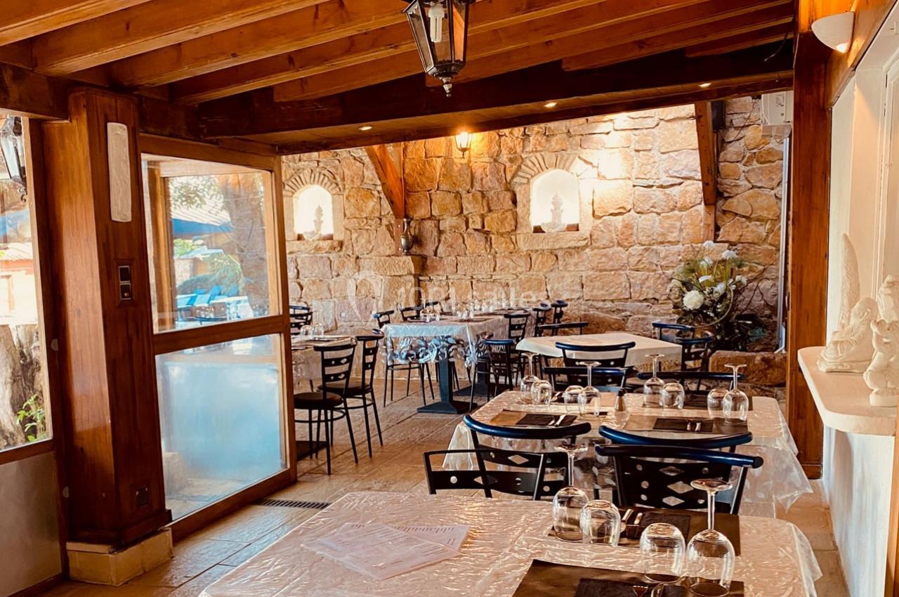 Salle de restaurant lumineuse avec tables dressées, murs en pierre et poutres apparentes en bois.