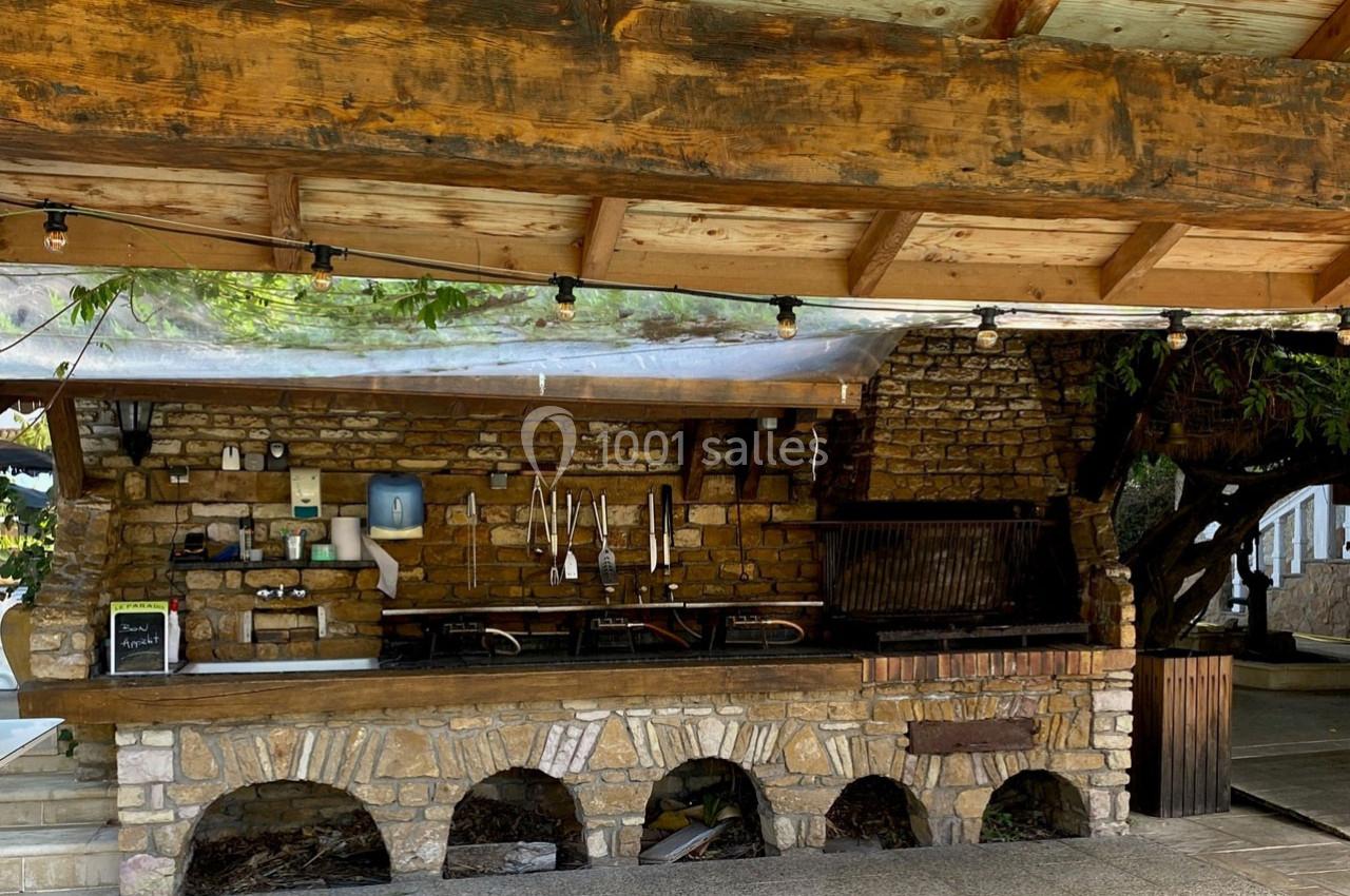 Cuisine extérieure en pierre sous un abri en bois, équipée de plaques de cuisson, ustensiles suspendus et éclairage.