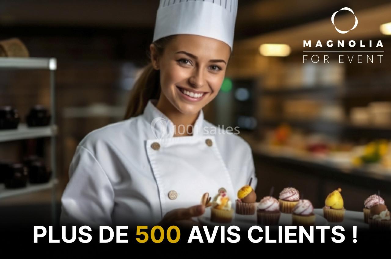 Une pâtissière souriante en tenue blanche présente un plateau de petits gâteaux dans une cuisine professionnelle.
