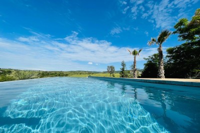 Piscine à débordement entourée de transats colorés sur une terrasse en bois, avec vue sur une campagne vallonnée.