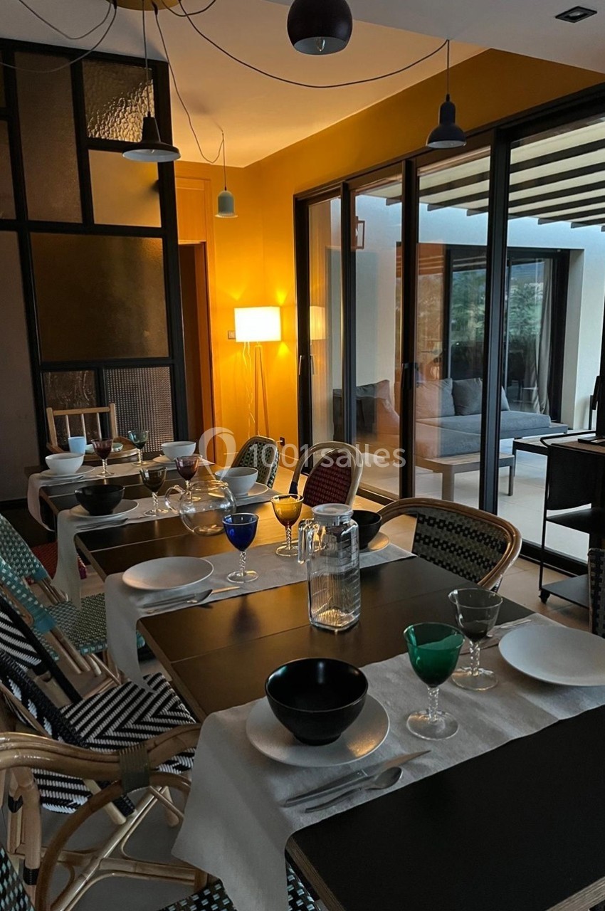 Salle à manger moderne avec table dressée, chaises en rotin, éclairage chaleureux et vue sur une terrasse vitrée.