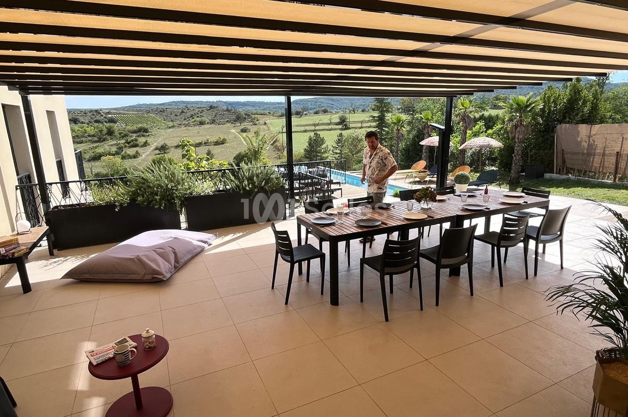 Terrasse couverte avec table dressée, vue sur jardin, piscine et paysage vallonné en arrière-plan.