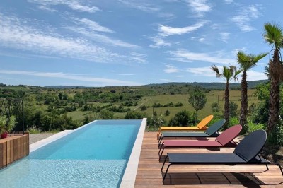 Piscine à débordement entourée de transats colorés sur une terrasse en bois, avec vue sur une campagne vallonnée.