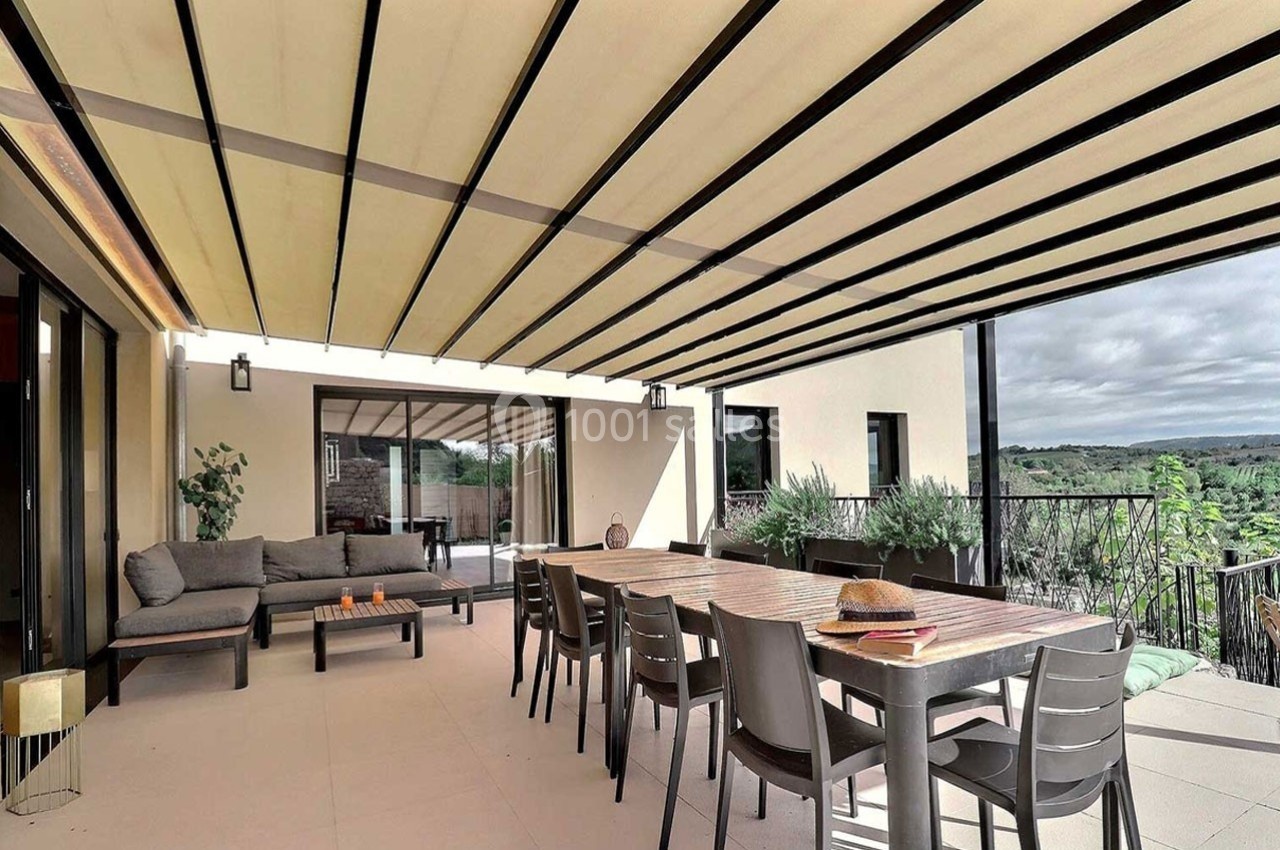 Terrasse spacieuse avec table, chaises, canapé et pergola à lames orientables, vue sur un paysage verdoyant.