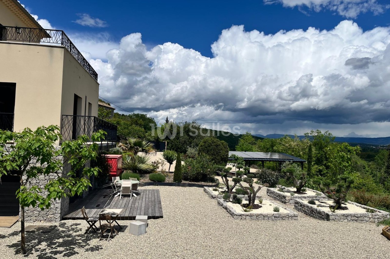 Terrasse de gravier aménagée avec un deck en bois, des massifs de plantes et d'arbustes taillés, une pergola à toiture solaire et une vue sur les collines sous un ciel nuageux.