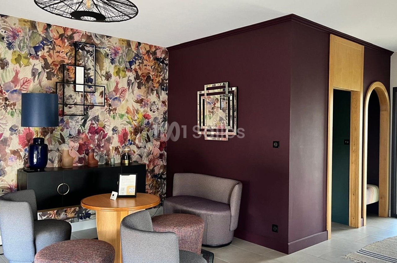 Salon au décor contemporain : papier peint fleuri multicolore sur un mur, mur bordeaux, fauteuils gris, table ronde en bois et petit meuble noir, encadrement d'une porte en bois clair. Let me check length: roughly 165 characters, too long. Need ≤125.