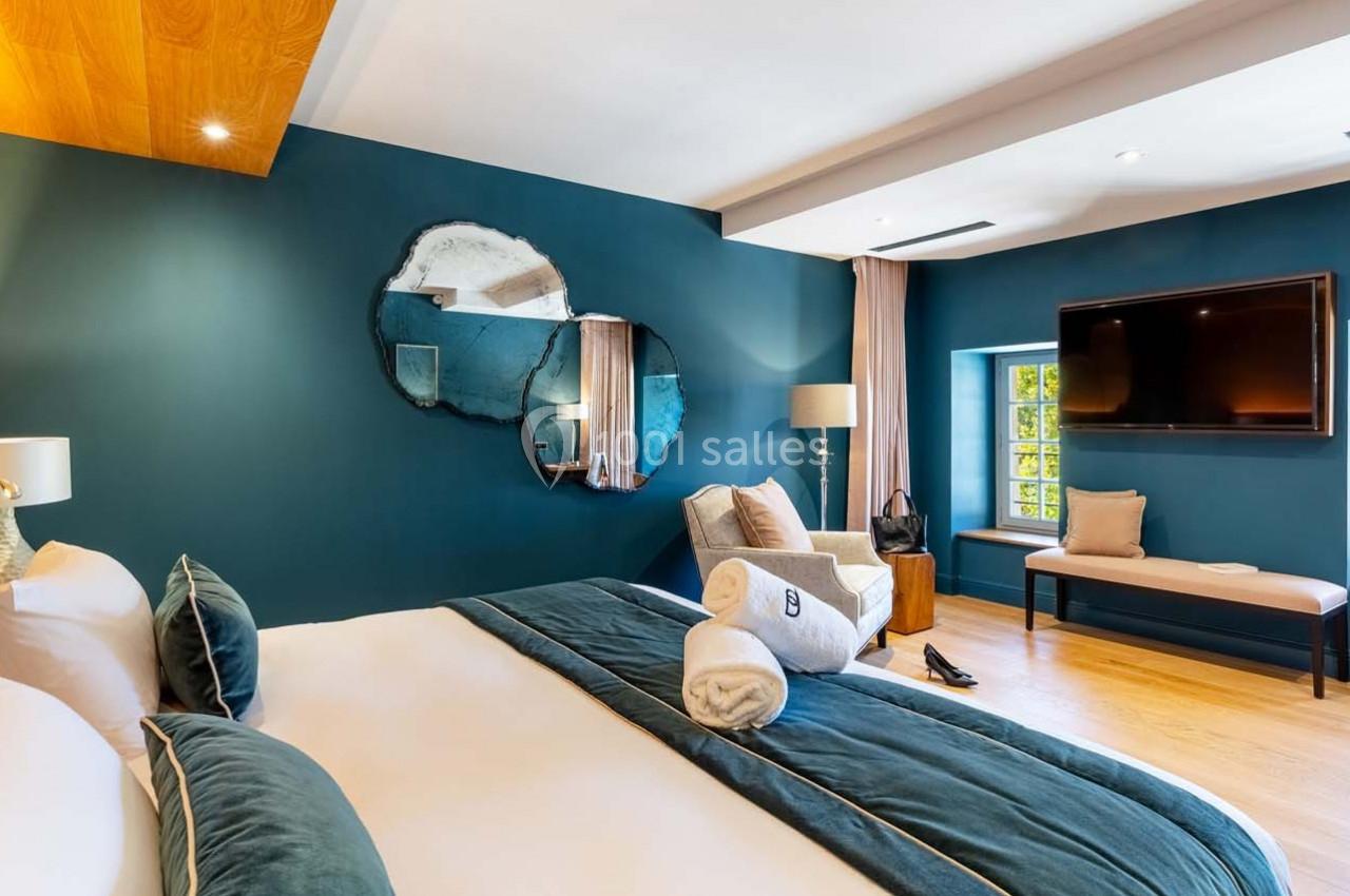 Chambre lumineuse avec lit double, murs bleu foncé, miroir décoratif, télévision murale et fauteuil près d'une fenêtre.