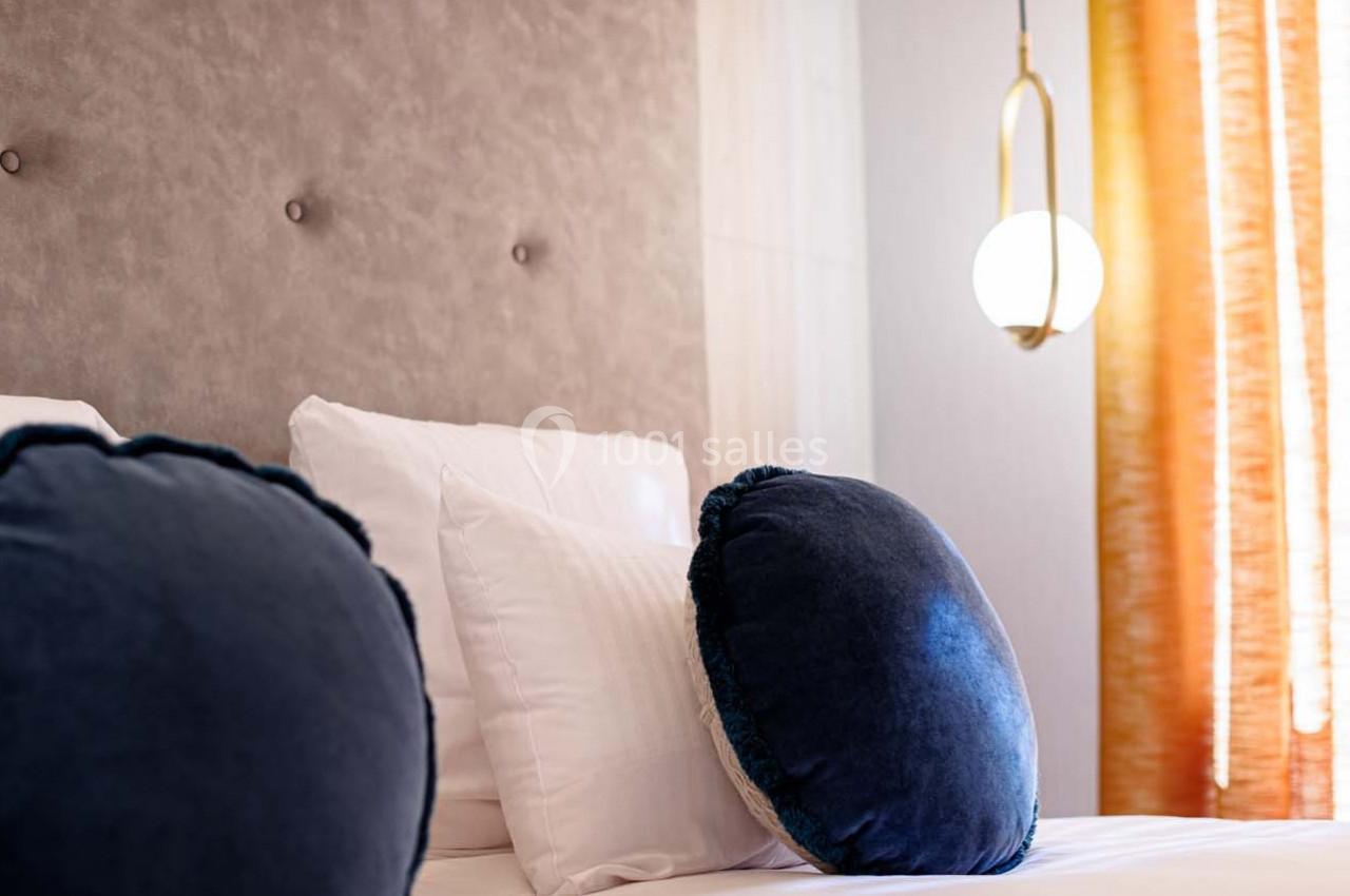 Lit avec coussins ronds bleu foncé, tête de lit capitonnée beige et rideaux orange laissant passer la lumière.