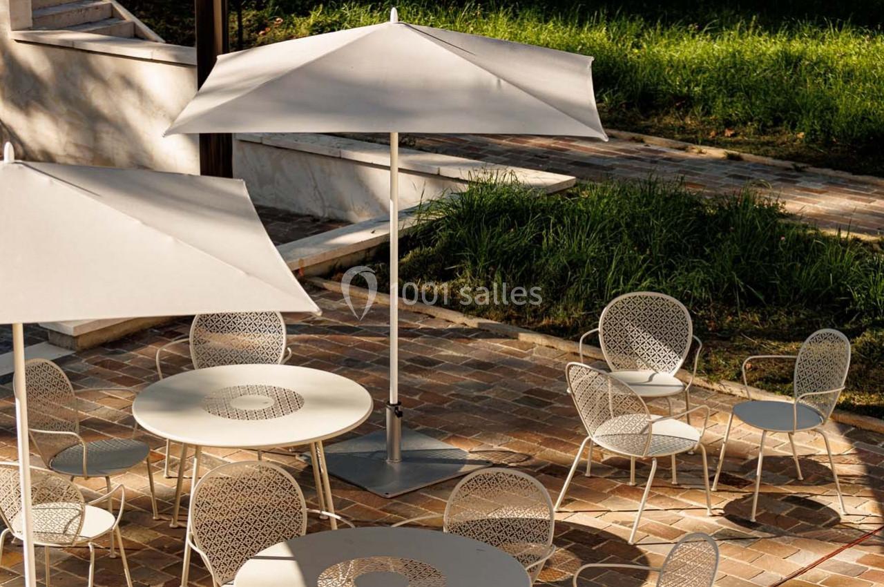 Tables et chaises blanches avec parasols sur une terrasse en pavés, entourée de verdure.
