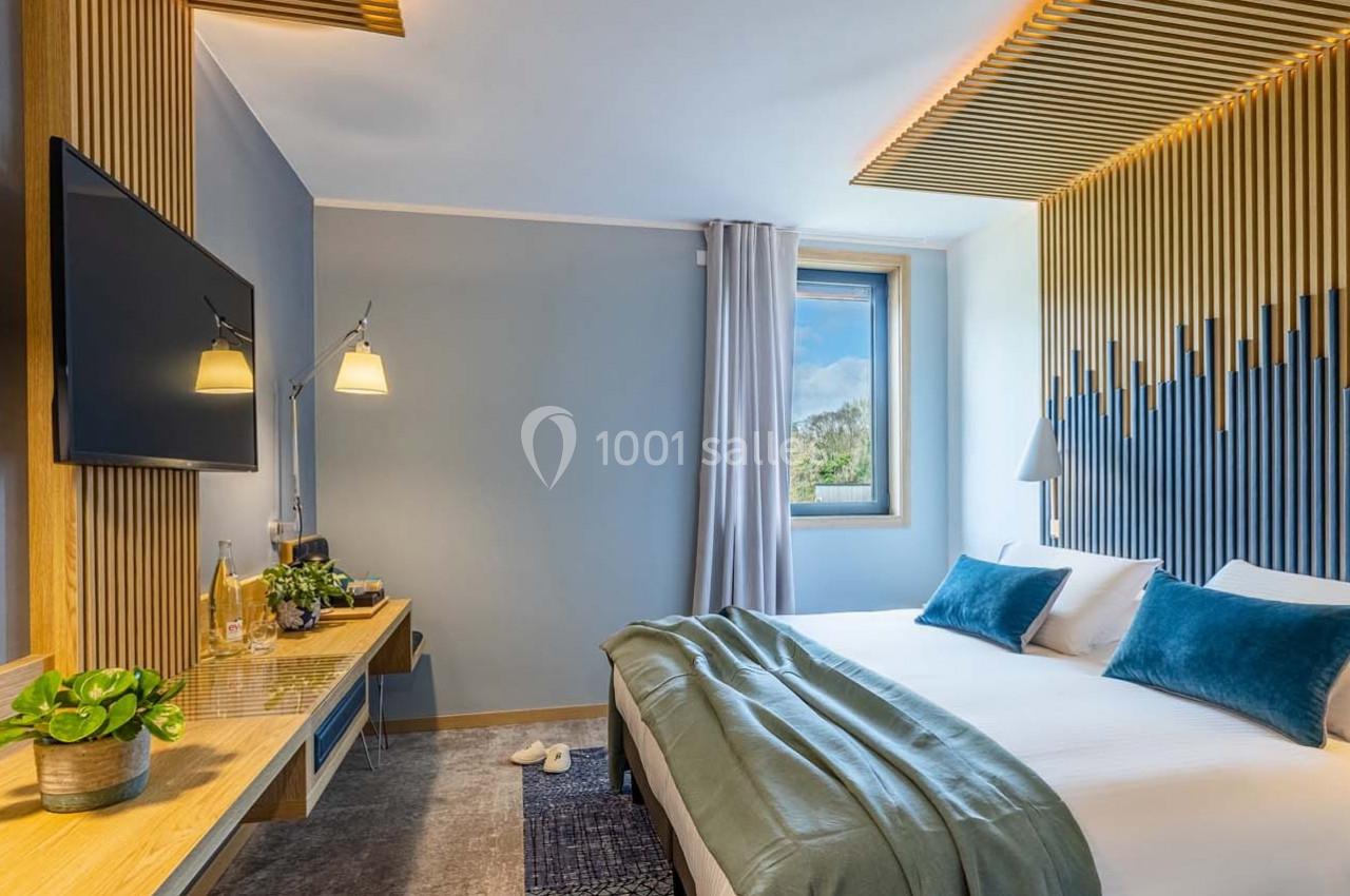 Chambre moderne avec lit double, décoration en bois, télévision murale, bureau et fenêtre donnant sur un paysage.